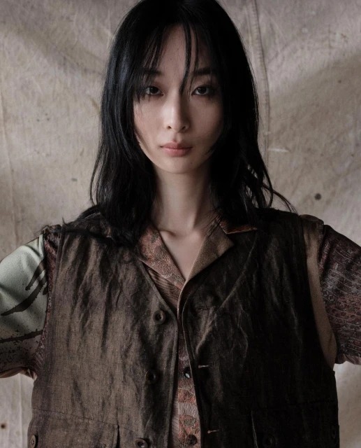 Portrait of a young Asian woman in a brown vest and patterned shirt against a textured backdrop, looking directly at the camera.