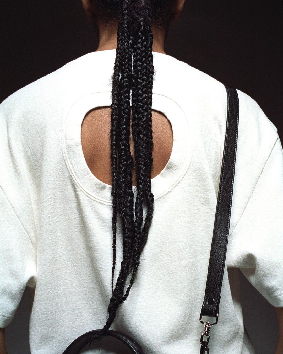 Person from the back wearing a white shirt with a circular opening and a black cross-body bag with braided hair against a dark background.