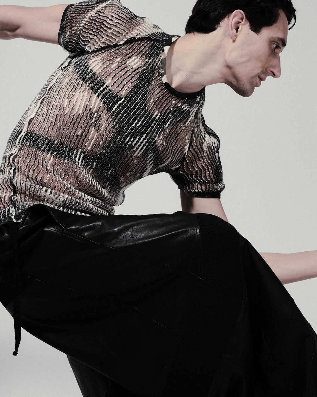 Fashion model wearing a patterned top and black leather skirt, striking a pose against a neutral backdrop in a studio setting.