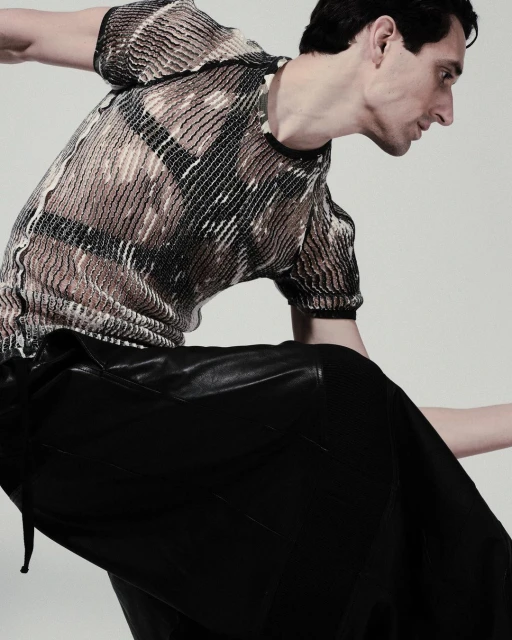 Fashion model wearing a patterned top and black leather skirt, striking a pose against a neutral backdrop in a studio setting.