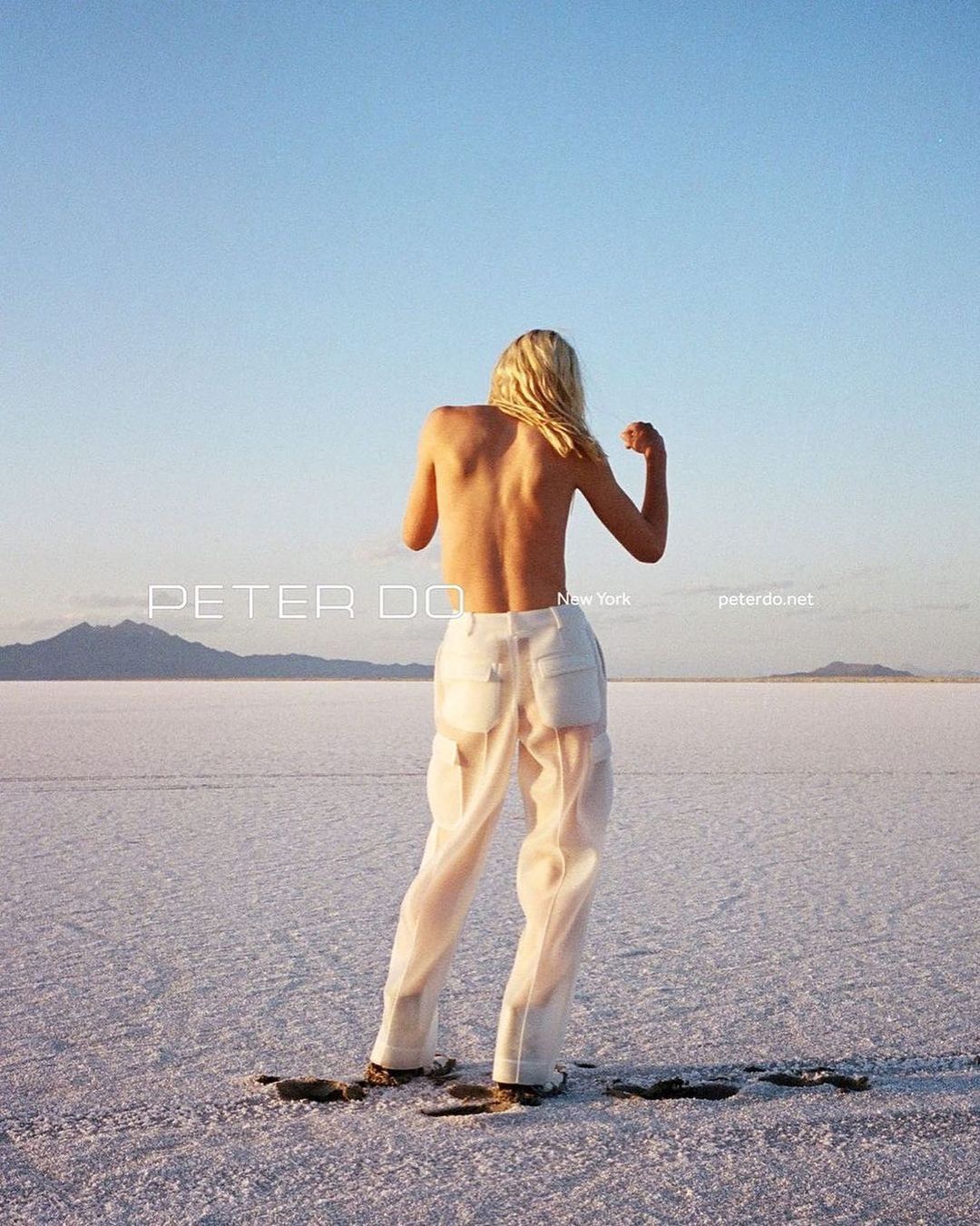 Blonde human stands in sheer white pants with mountain backdrop under blue sky and sunlit landscape of what appears to be salt flats.