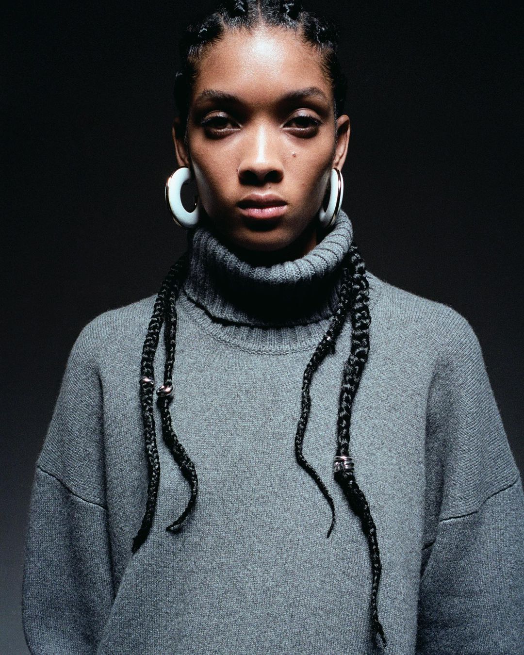 Close-up studio portrait of an adult female with braided hair wearing a grey turtleneck sweater and hoop earrings against a dark background.