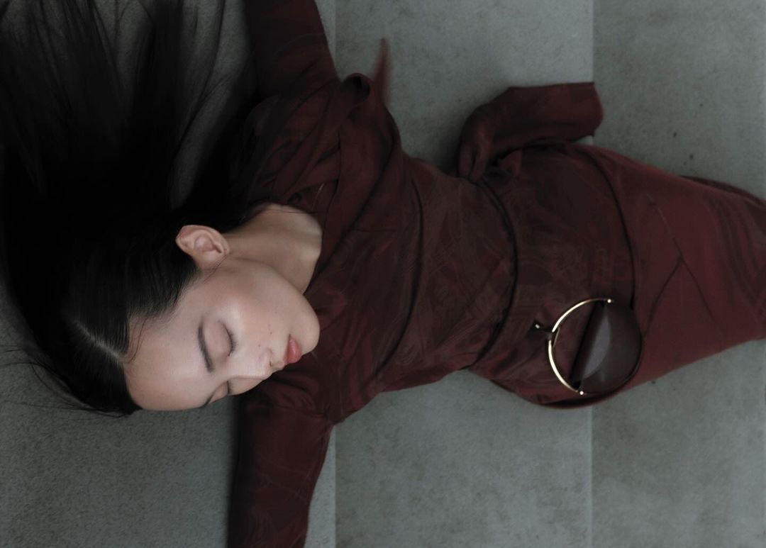 A woman in a maroon dress lies on a gray surface with her eyes closed and black hair spread out, accessorized with round sunglasses.