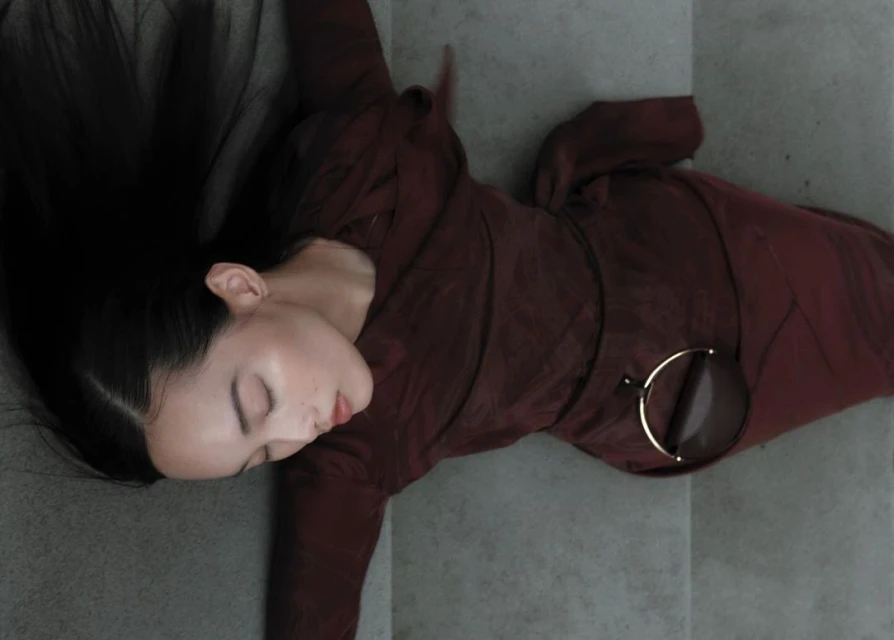 A woman in a maroon dress lies on a gray surface with her eyes closed and black hair spread out, accessorized with round sunglasses.
