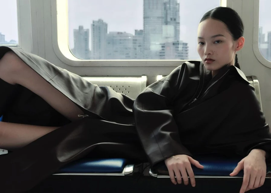 A young, dark-haired woman lies on blue train seats wearing a dark, oversized jacket, with a modern city visible through the window.