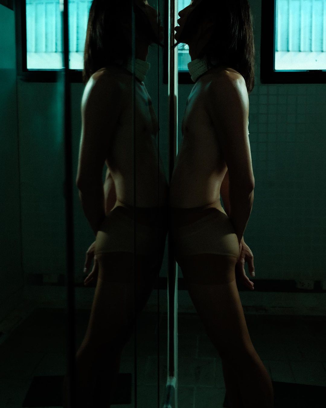 A person stands silhouetted in front of a mirror in a dark room, showcasing the reflection of their body in a minimalist, intimate composition.