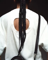 Close-up back view of a person wearing a white t-shirt with a round back cut-out, black braided hair, and a black strap bag.