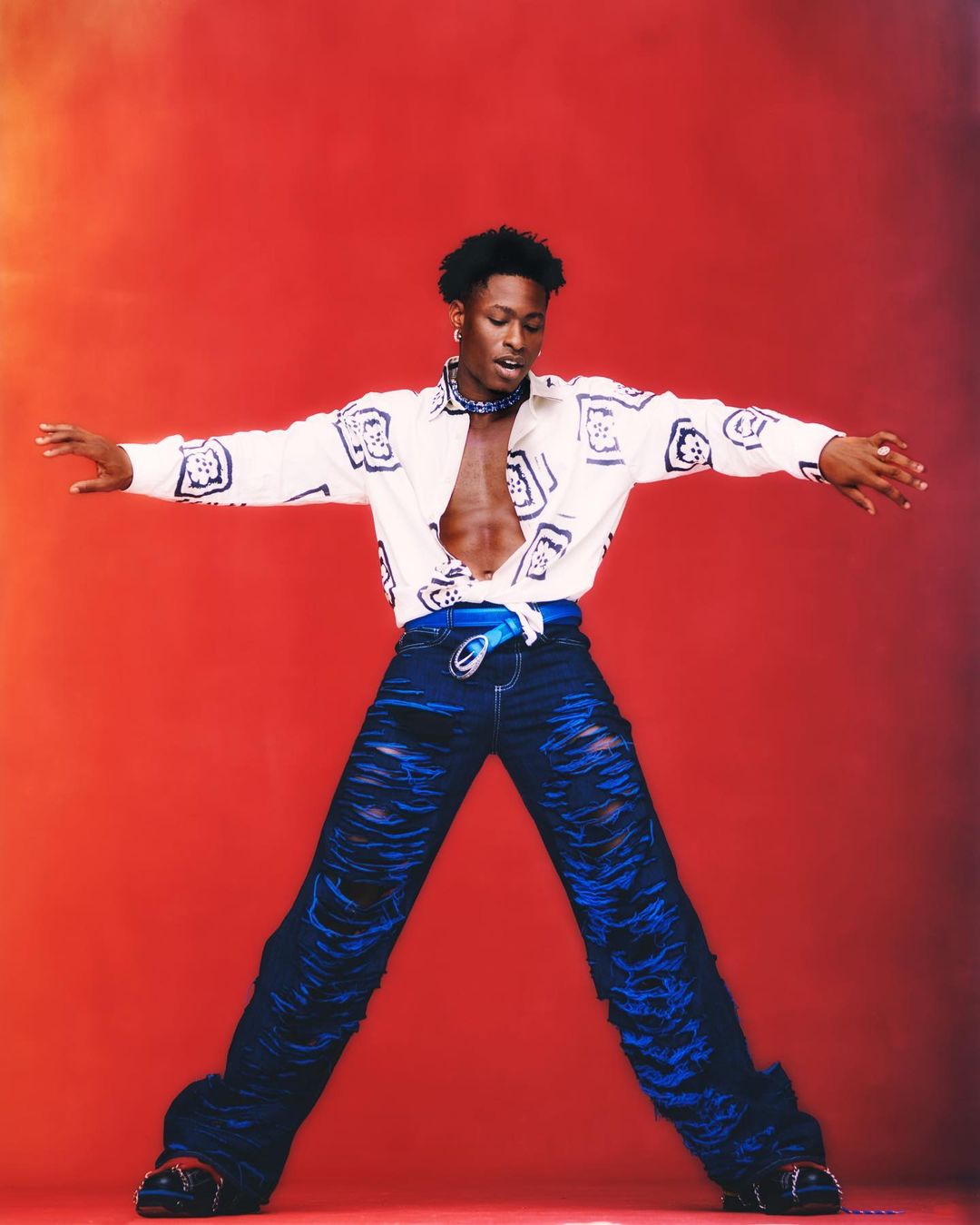 Stylish adult male with open shirt and ripped jeans poses with arms outstretched against a vibrant red studio background.