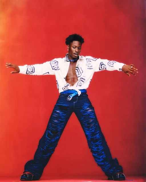 Stylish adult male with open shirt and ripped jeans poses with arms outstretched against a vibrant red studio background.