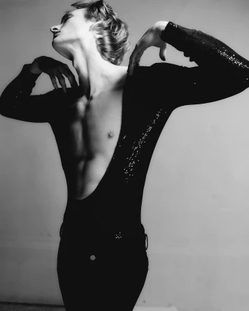 Monochrome portrait of a male model posing dramatically with arched back in a black long-sleeve top against a grey seamless background.