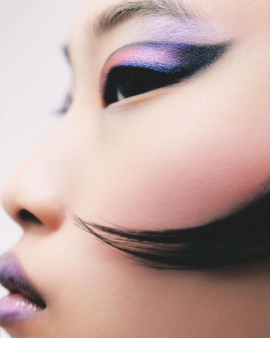 Close-up shot of a woman's face showing striking makeup including purple lipstick and iridescent eyeshadow with long, dramatic eyelashes.
