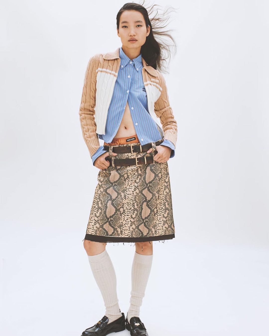 Full shot of a young adult woman in a fashion pose, wearing a striped shirt, brown and white cardigan, and a snakeskin-print skirt on a white background.