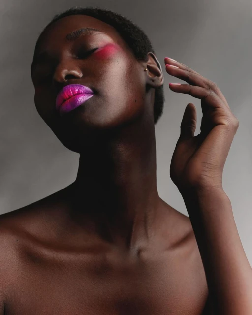 Closeup portrait of Black woman with eyes closed, vivid magenta and pink makeup on lips and cheek, against a neutral gray background.