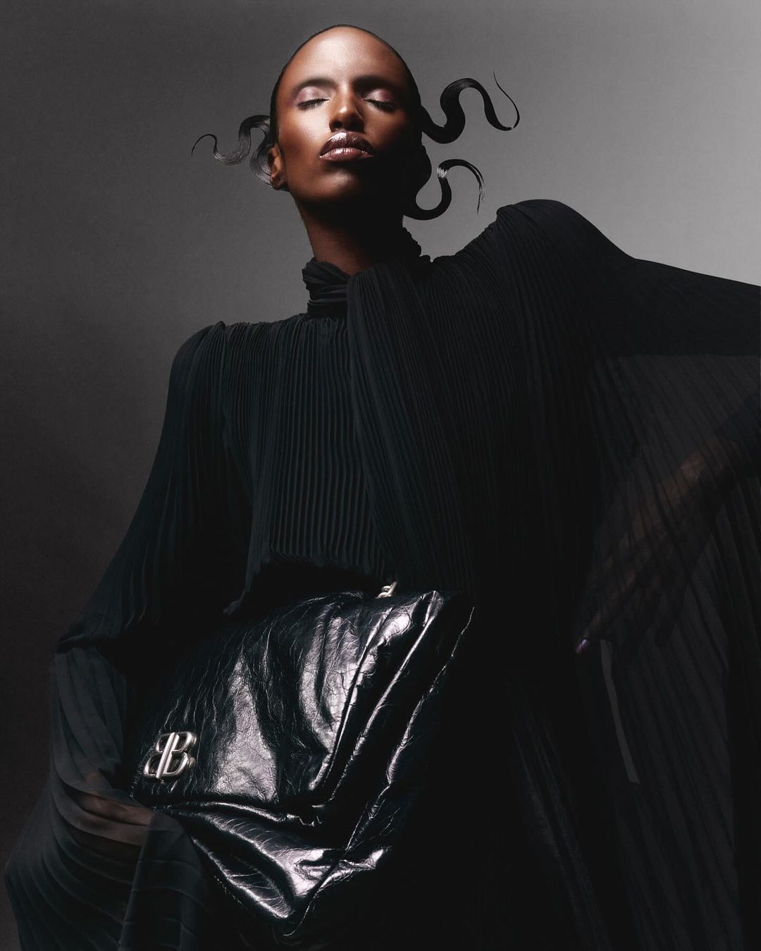 Fashion shot of Black female model in draped black dress, dramatic pose, and black bag with logo against a grey background in studio lighting.