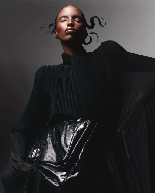 Fashion shot of Black female model in draped black dress, dramatic pose, and black bag with logo against a grey background in studio lighting.