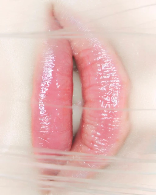 Close-up of pursed pink lips between clear plastic wrap, creating a distorted effect and highlighting the texture and color.