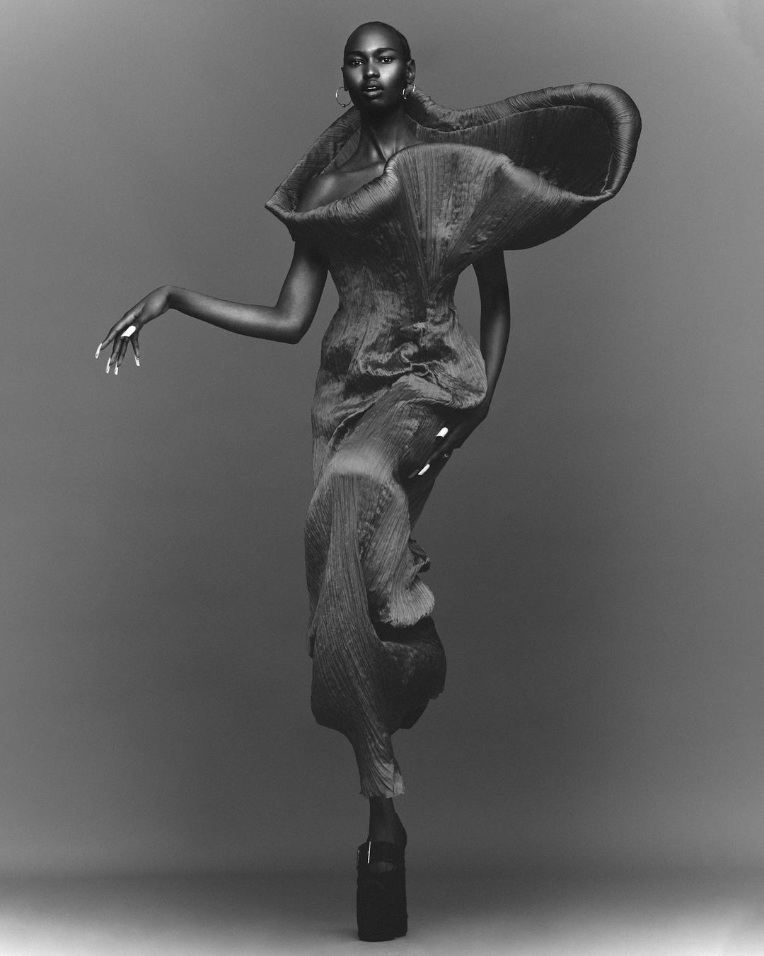 A tall, thin, African female fashion model stands in a sculpted dark gown and platform shoes against a grey studio backdrop.