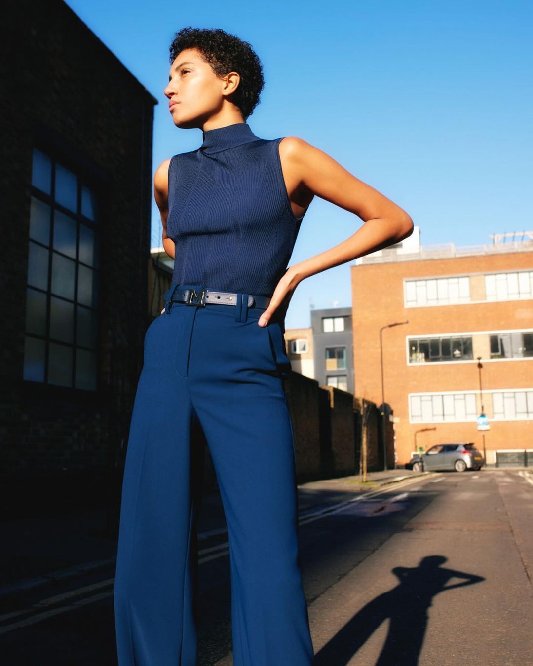 Adult woman in a blue ribbed top and flared pants stands outside a brick building on a street on a sunny day. 