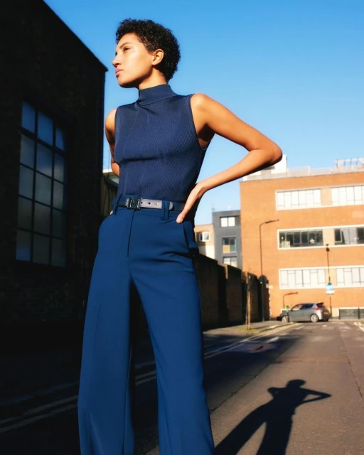 Adult woman in a blue ribbed top and flared pants stands outside a brick building on a street on a sunny day. 