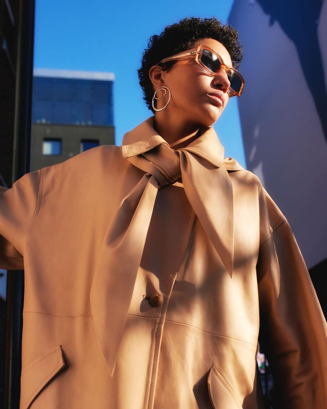 Elegant woman in tan coat and sunglasses in urban setting. Natural lighting highlights textures and architectural background.