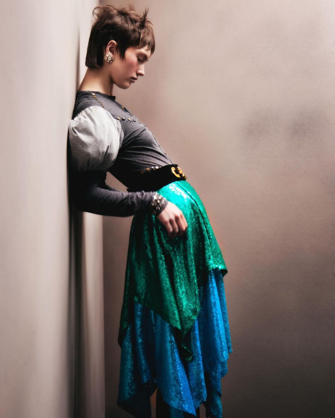Fashion shot of an adult female in a gray top and colorful, layered sequin skirt, leaning against a wall in a photo studio.