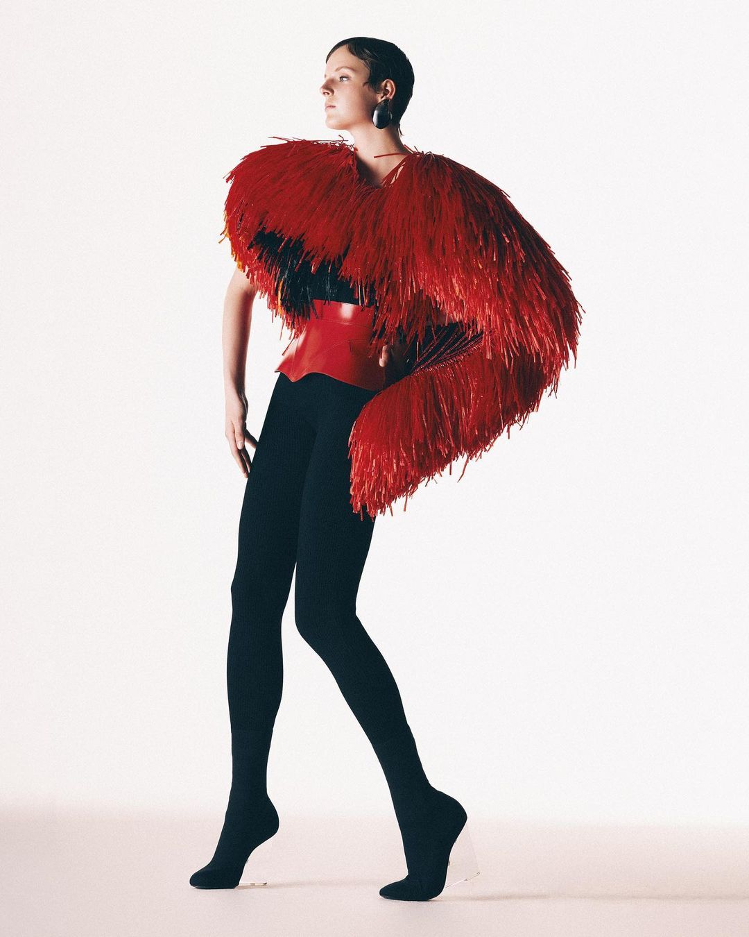Full-body shot of a female model in a red feather boa and black leggings, posing in a bright white studio setting. Fashion editorial.
