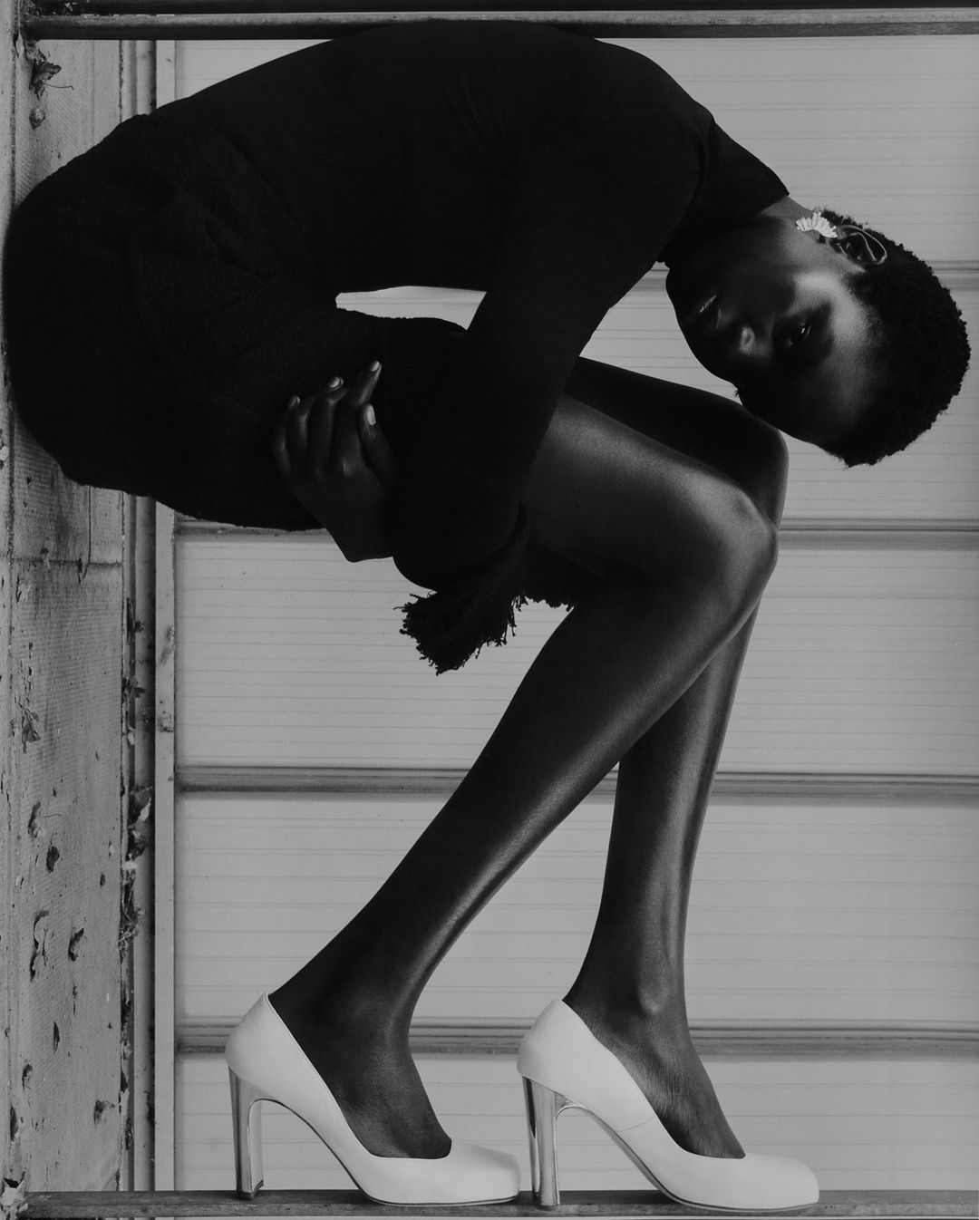 Monochrome portrait of an African female in dark dress and white high heels, leaning on a textured wall in a fashion pose.