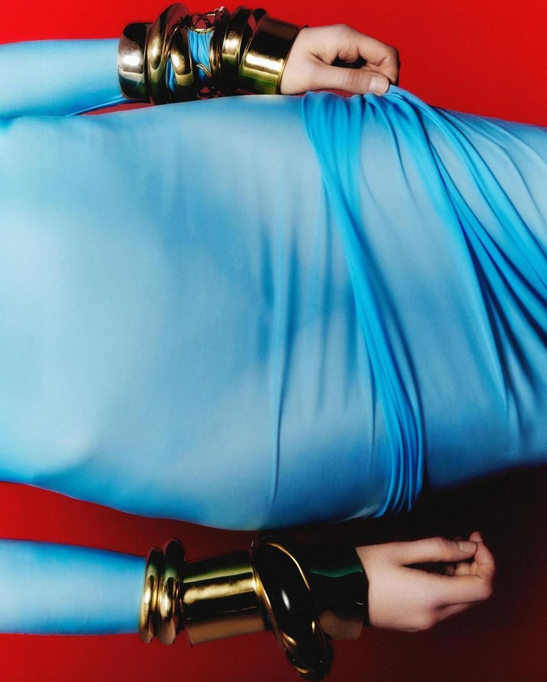 Close-up of a person in a light blue dress with gold bracelets against a red backdrop, highlighting fashion and style.