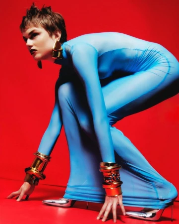 Fashion photo of a model in a full-body blue bodysuit and statement jewelry posing in a crouch in a bright red studio setting.