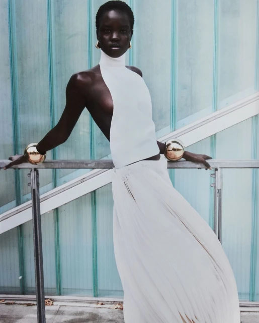 Fashion portrait of a slim African-American adult female wearing a white dress and gold bracelets, posing in front of a glass panel wall.