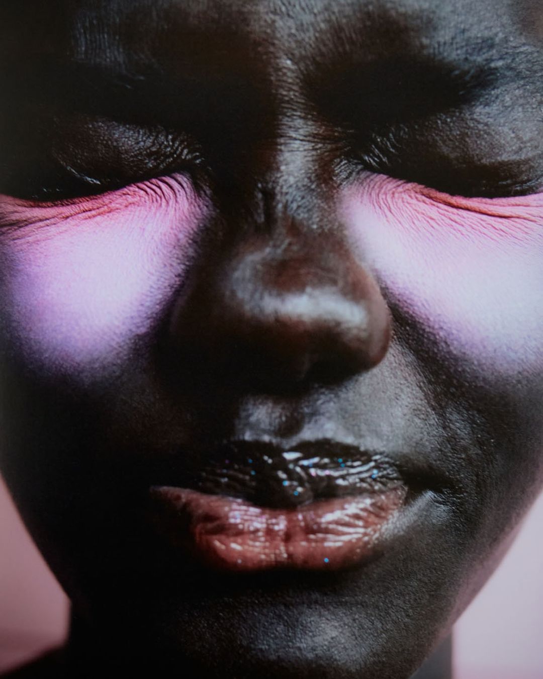 Portrait of a dark-skinned woman with heavy pink eye makeup and glossy lips against a soft pink background, photographed with a close-up framing.