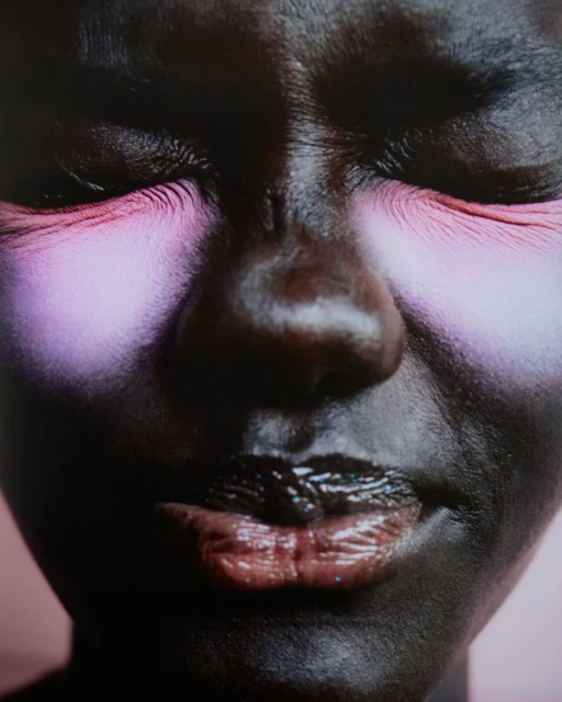 Portrait of a dark-skinned woman with heavy pink eye makeup and glossy lips against a soft pink background, photographed with a close-up framing.