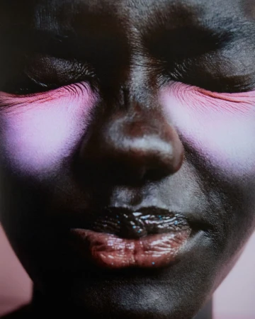 Portrait of a dark-skinned woman with heavy pink eye makeup and glossy lips against a soft pink background, photographed with a close-up framing.