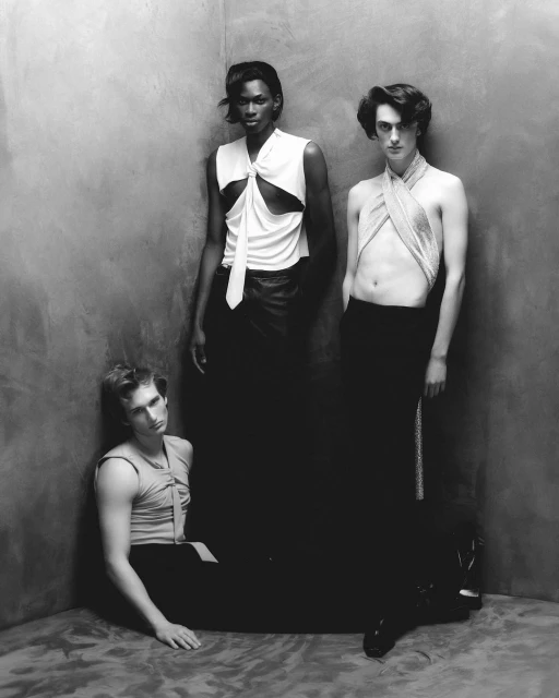 Three fashion models pose in black and white, wearing draped tops and dark pants, against a textured wall in a studio environment.