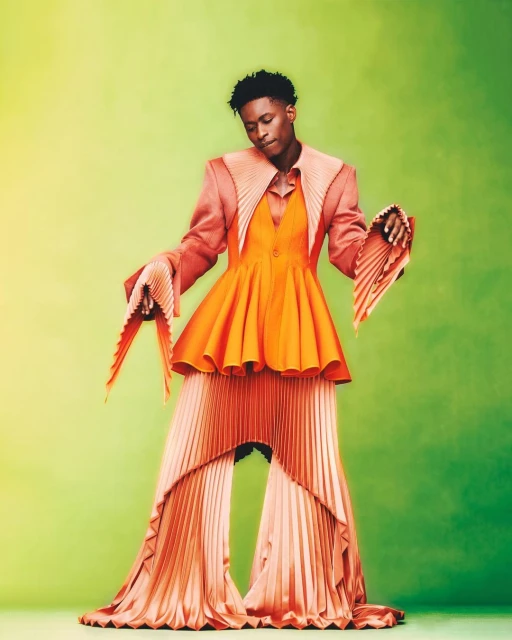 Young man in orange pleated fashion against vibrant green studio backdrop. Statement sleeves, blazer, and pants create a bold, stylish look.