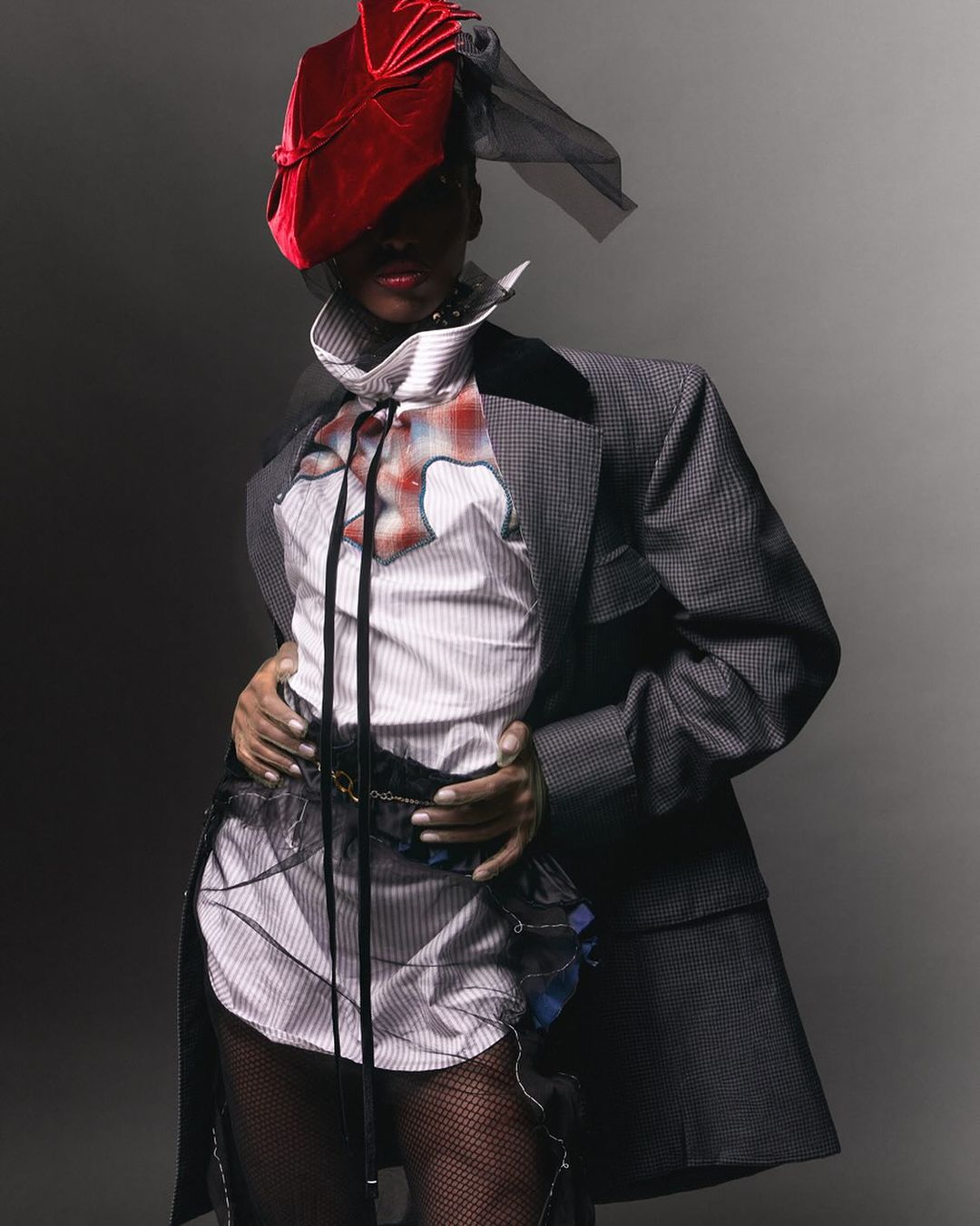 Fashionable individual in a tailored grey suit, unique hat, and stylish shirt, posing in a studio setting for a high-fashion editorial shot.