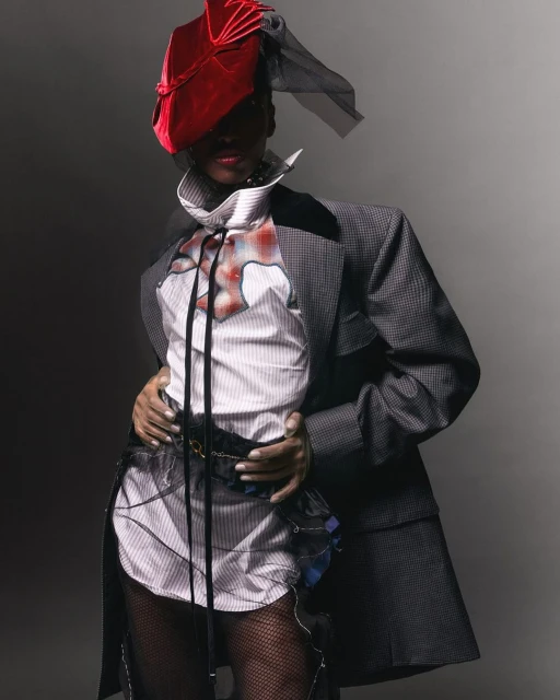 Fashionable individual in a tailored grey suit, unique hat, and stylish shirt, posing in a studio setting for a high-fashion editorial shot.