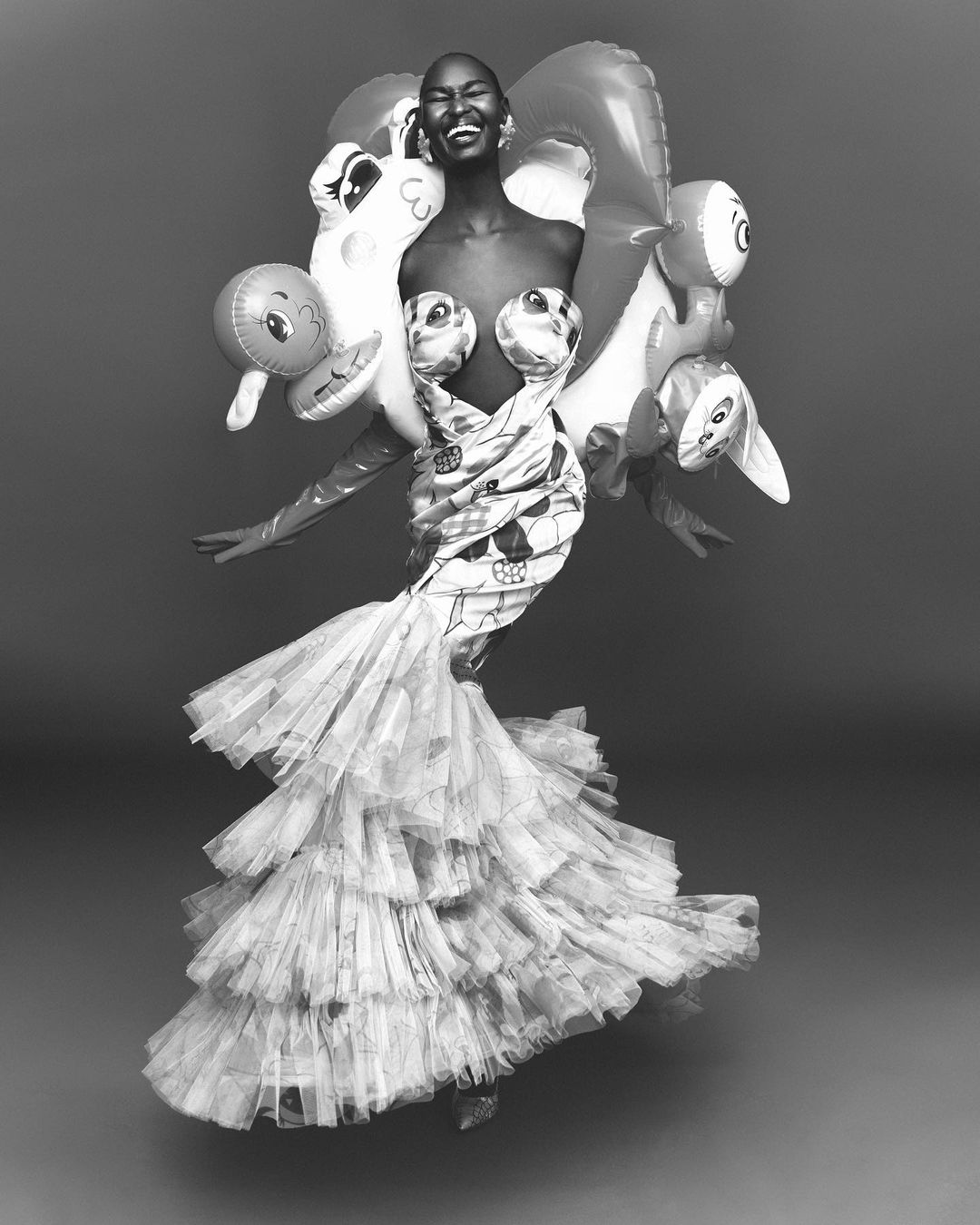 Elegant Black woman in couture gown made from maps and toy inflatables, striking a playful pose against a neutral gray backdrop.