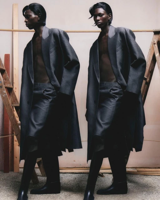 Two men pose in oversized grey coats and black boots in a studio setting with a ladder and neutral-toned backdrop for a fashion editorial.