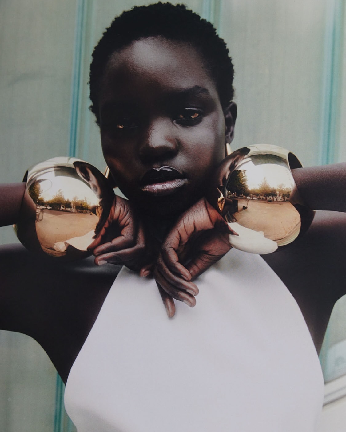 Close-up of a black model with short hair wearing a white top and large gold bracelets, hands crossed, posing in front of a striped wall.