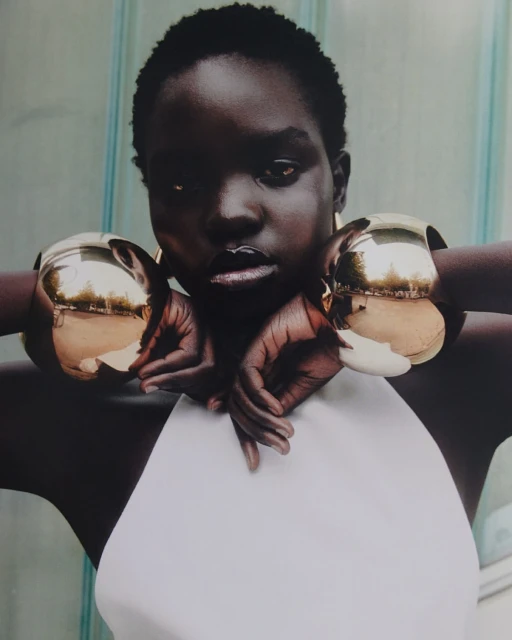 Close-up of a black model with short hair wearing a white top and large gold bracelets, hands crossed, posing in front of a striped wall.