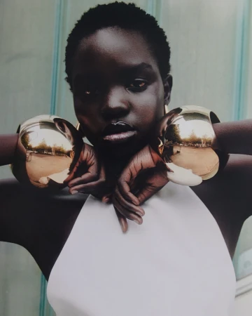 Close-up of a black model with short hair wearing a white top and large gold bracelets, hands crossed, posing in front of a striped wall.