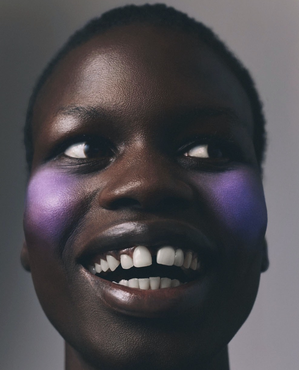 Close-up studio portrait of smiling African woman with gap teeth, wearing purple makeup on cheekbones, against a grey backdrop.