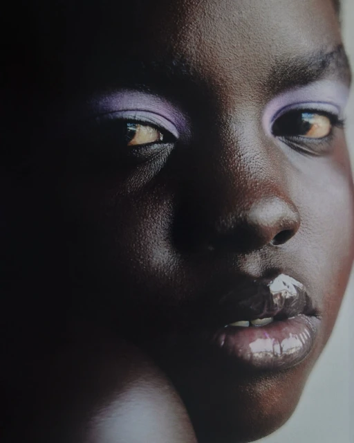 Close-up portrait of a dark-skinned woman with soft, diffused lighting and glossy lips, featuring lilac eyeshadow and a dark background.