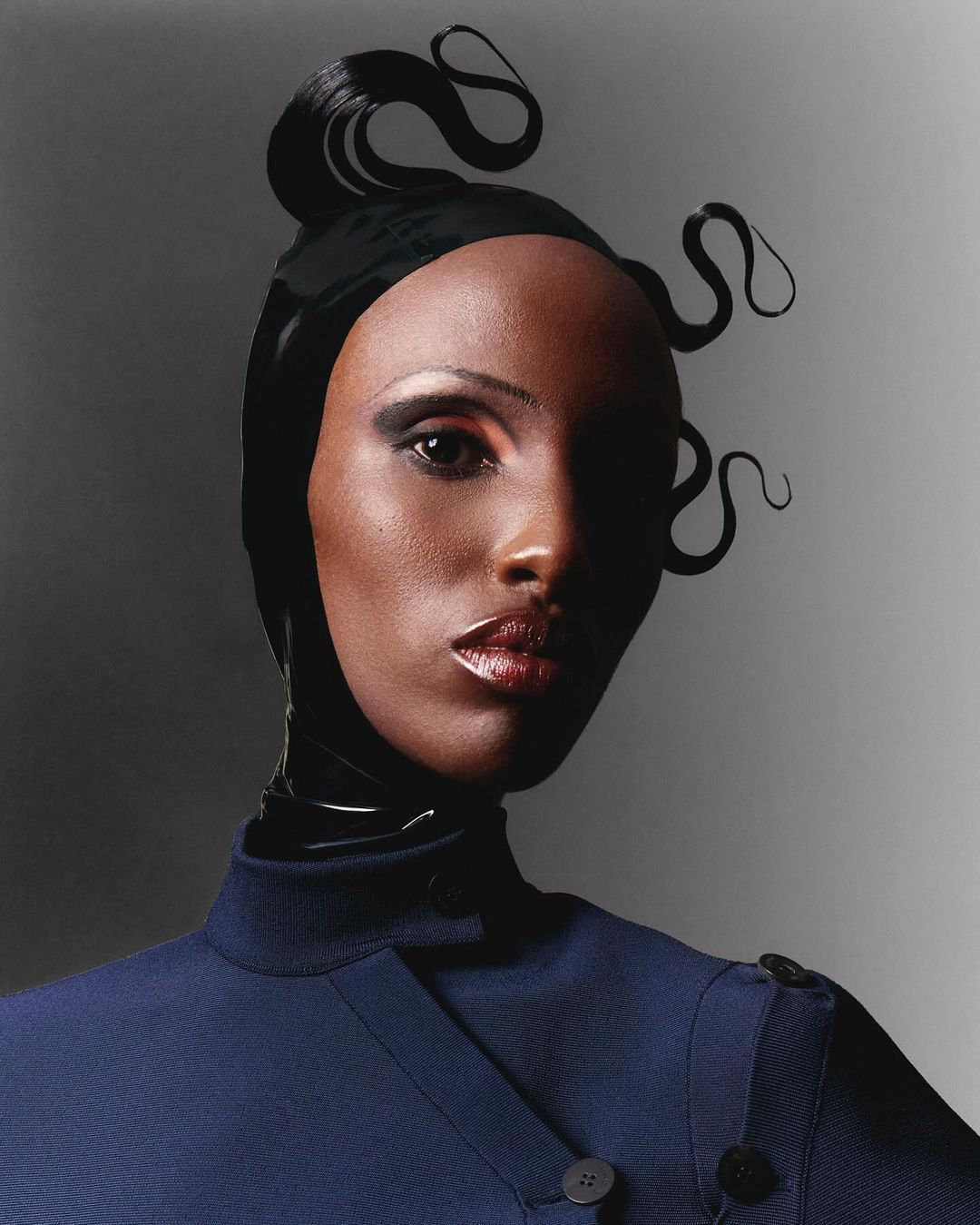 Headshot of a dark-skinned woman with stylized black hair wearing a navy coat against a gray background in a fashion portrait.