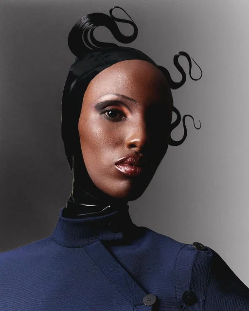Headshot of a dark-skinned woman with stylized black hair wearing a navy coat against a gray background in a fashion portrait.