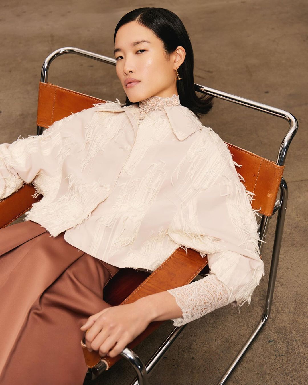 Elegant Asian woman sits in a modern chair, wearing a pale jacket and copper pants. Studio lighting, clean background, and a relaxed pose.