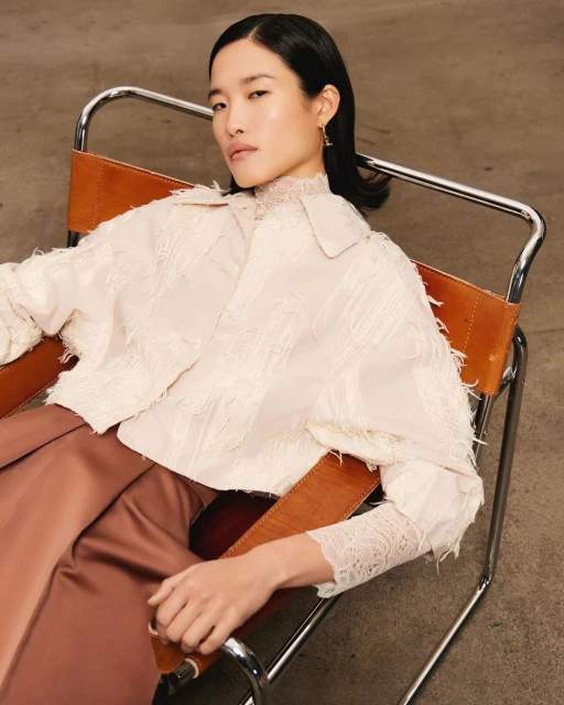 Elegant Asian woman sits in a modern chair, wearing a pale jacket and copper pants. Studio lighting, clean background, and a relaxed pose.
