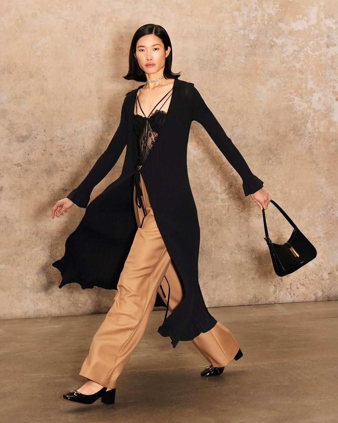 A stylish Asian woman strides confidently in a long black cardigan, wide-leg trousers, and heels, carrying a black handbag against a textured backdrop.