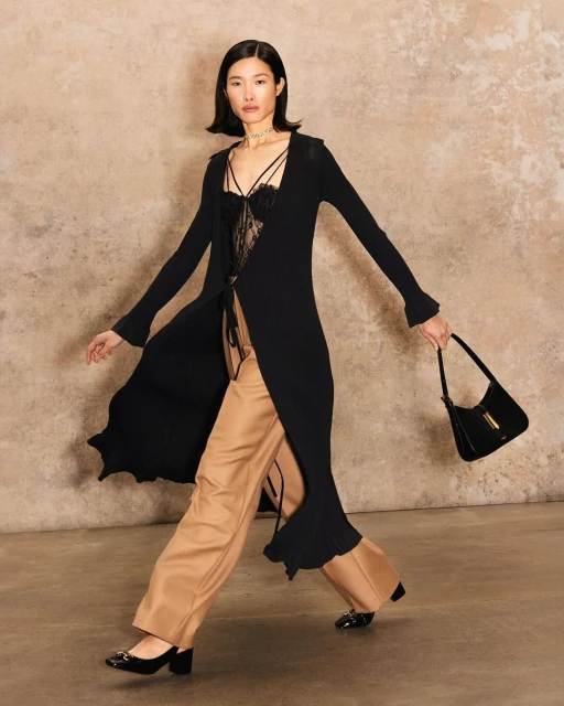 A stylish Asian woman strides confidently in a long black cardigan, wide-leg trousers, and heels, carrying a black handbag against a textured backdrop.